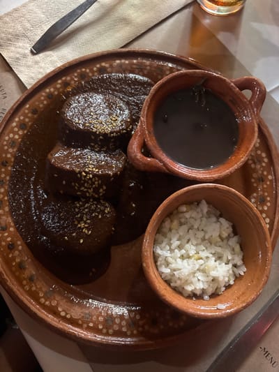The image displays a meal served on a terracotta plate, presumably in a restaurant located in San Sebastián Xolalpa, Mexico. The central item is a large, round, reddish-brown earthenware plate with a textured, dotted rim. On this plate, three distinct, cylindrical portions of dark brown mole, heavily garnished with white sesame seeds, are arranged. To the right of the mole, a small, handled terracotta cup contains a dark, smooth liquid, consistent with black bean soup or a dark sauce. Below this, a smaller, open terracotta bowl holds white rice, which appears to have a few light yellow specks mixed in.
In the upper left corner of the frame, a beige, textured paper placemat or napkin is visible, with the handle of a silver-colored utensil, likely a knife, resting upon it. A translucent glass containing a yellow-amber liquid is partially visible in the upper right background, suggesting a beverage such as beer. A white surface, possibly a tablecloth, is visible beneath the placemat and the glass. The corner of a paper item, with the word "MENU" partially discernible, is visible in the lower right. The scene is illuminated by warm, ambient lighting. No individuals are visible in the frame.