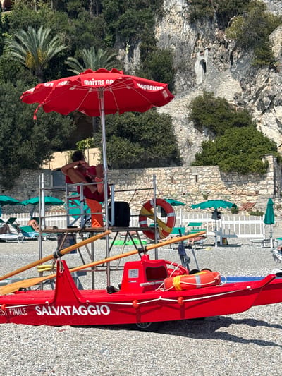 The image depicts a sunny day at a beach in Finale Ligure, Italy, characterized by its pebble shore and rocky cliffs. In the foreground, a bright red lifeguard boat, labeled "FINALE SALVATAGGIO," is prominently positioned. Two oars are visible attached to the boat's structure. Above and behind the boat, a lifeguard sits on a raised platform under a large red umbrella displaying the text "MAITRE NAD" and "OGORRIST." The lifeguard is wearing a red shirt and appears to be relaxed, with their legs crossed. Further back, a white picket fence separates the beach from a landscaped area with green beach umbrellas and lounge chairs, some occupied by sunbathers. The background features lush green vegetation cascading down a steep, rugged cliff face, with remnants of ancient stone structures integrated into the rock. The overall scene suggests a typical summer day at a well-equipped beach resort.