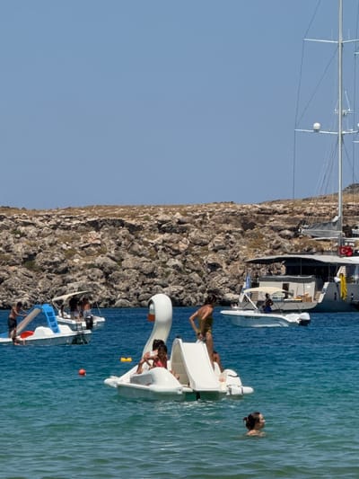 In the waters of Lindos, Greece, on a clear day, several boats and people are visible. In the foreground, a white swan-shaped paddle boat has at least two individuals on it, one appearing to be a child and the other an adult. The paddle boat has a slide attached to it. To the left of the swan boat is a smaller white boat with a blue slide, carrying several people. A man in dark shorts stands on the shore of this boat. Further in the background, a larger white yacht with multiple masts is moored. Another smaller, darker boat with a canopy is anchored nearby, displaying a Greek flag and a Turkish flag. The water is a vibrant turquoise, and a rocky, arid hillside forms the backdrop. A woman is swimming in the water in the lower right quadrant of the image.