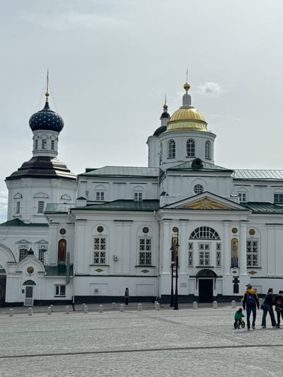 The image depicts the Resurrection Cathedral in Arzamas, Russia. The cathedral, characterized by white walls and green roofs, exhibits intricate architectural details including various windows and golden embellishments. People are visible along the right side of the building. The foreground shows a paved walkway. The sky is overcast.