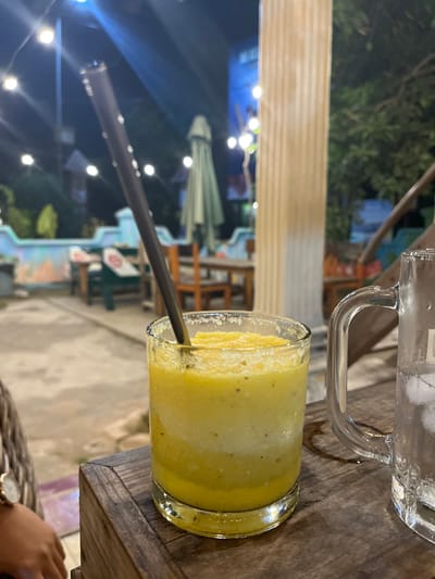 The image shows a close-up of a glass of yellow smoothie or juice with a dark straw in it, resting on a wooden table. Behind the smoothie, a glass mug filled with ice and liquid can be seen. The scene appears to be outdoors, with blurred background elements suggesting an evening setting in Karimunjawa, Indonesia. String lights are visible in the background, indicating it might be nighttime. The overall ambiance suggests a relaxed, possibly tropical, evening at an eatery or bar. The focus is on the drinks, with the background providing context without being sharply defined. The composition and lighting suggest a casual, candid moment.