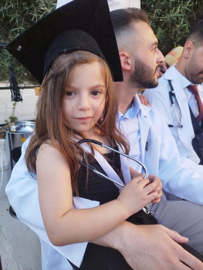 This image captures an outdoor scene in Al-Khalil, Palestinian Territory, featuring a young girl in the foreground with two adult males. A light-skinned girl, estimated 4-7 years old, with long brown hair, wears a black academic mortarboard with a tassel and a black top overlaid with white straps resembling a small lab coat. She holds a silver and black stethoscope across her chest, looking straight ahead. She is seated on the lap of a light-skinned adult male, likely in his 20s-30s, with a beard, wearing a light blue shirt under a white lab coat; his right arm is around the girl, and he looks to his right. To his right, a second light-skinned adult male, similarly aged and dressed in a white lab coat over a dark vest and red tie, with a stethoscope around his neck, is partially visible and also looking right. The background features blurred green foliage and a glimpse of a metallic container on the left, with a yellow object displaying partial Arabic script (e.g., "على") in the distant right.