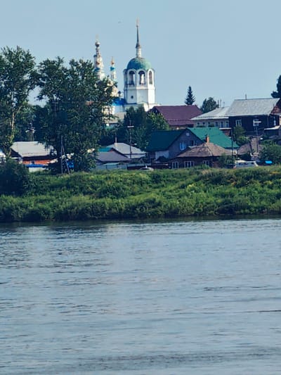 The image depicts a view of the city of Yeniseysk, Russia, from across a body of water. In the background, a prominent church with a blue-green dome and a tall white steeple stands out against a clear sky. Several other buildings with pitched roofs, some appearing to be houses with chimneys, are visible clustered together. Lush green trees and vegetation line the riverbank in the middle ground, with some smaller trees scattered amongst the buildings. The foreground is dominated by the rippling surface of a wide river, reflecting the light blue of the sky.