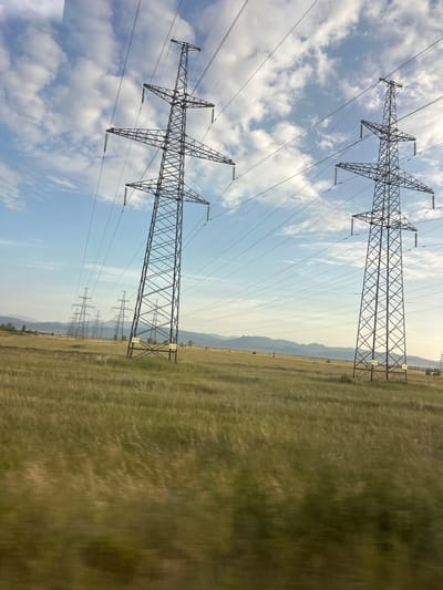 This image shows a series of electricity pylons traversing a vast, open landscape under a partly cloudy sky. The pylons, constructed of dark metal lattice, are a dominant feature, stretching across the frame and receding into the distance. They are spaced at intervals, with thick power lines strung between them. The pylons in the foreground are sharp and clear, while those further back become progressively less distinct, suggesting a significant distance.
The landscape is characterized by dry, golden-brown grass, indicating a rural or steppe-like environment. The grass appears somewhat blurred, suggesting movement, perhaps due to wind or the viewpoint being in motion. In the background, a range of low hills or mountains is visible, fading into a hazy horizon. The sky is a pale blue, dotted with scattered white clouds, some of which are quite fluffy.
The lighting suggests it is daytime, likely late morning or afternoon, given the angle of the light and the brightness of the scene. The overall atmosphere is one of vastness and a sense of distance, with the industrial elements of the pylons juxtaposed against the natural, expansive landscape. There is no clear indication of people or any specific activity taking place, other than the presence of the power transmission infrastructure. Some of the pylons appear to have yellow rectangular signs attached to them, but the text is not legible. The location is identified as Novonikolaevka, Russia.