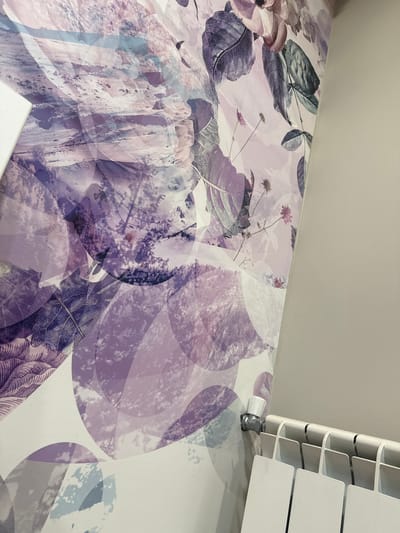 The image shows a close-up view of a radiator with a white control valve attached to a pipe, mounted on a wall with floral wallpaper. The wallpaper features abstract floral designs in shades of purple and pink, with some areas appearing translucent and layered. The radiator is white and appears to be a standard panel radiator. The wall to the right of the wallpaper is a neutral, light gray color. The scene is indoors and appears to be in a domestic setting. There are no people or animals visible in the image. The image does not contain any information that would allow for identification of its location within the city of Petkovo, Bulgaria.