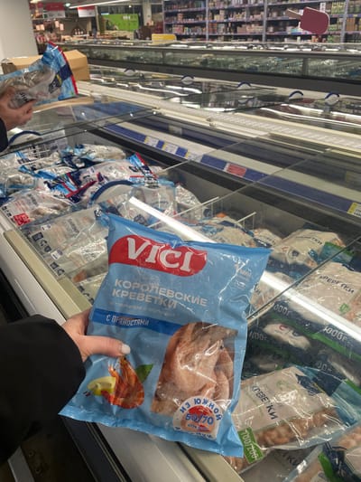 The image captures a close-up view inside a supermarket, specifically in the frozen food section. The main subject is a person's hand holding a light blue plastic bag of frozen shrimp from a display freezer. The hand is visible from the wrist down, clad in a dark sleeve, and shows fingernails painted a light, possibly white, color.
The bag prominently displays the brand name "VICI" in red lettering on a white background within a red rectangle. Below this, large Cyrillic text reads "КОРОЛЕВСКИЕ КРЕВЕТКИ" (Royal Shrimps). Further down, it specifies "в панцире с головой" (in shell with head) and "С ПРЯНОСТЯМИ" (with spices). The bag features an image of a cooked shrimp with a slice of lemon and a sprig of herb at the bottom right. A circular blue sticker on the bag contains more Cyrillic text, including "ЮЖНОЙ" (Southern) and "50/70 ШТУК" (50/70 pieces), indicating the size or count of the shrimp. The product inside the bag is visible through a clear window, showing frozen, pale pink shrimp. Frost and condensation are visible on the outside of the bag.
The background shows the interior of a refrigerated display case, filled with numerous other bags of frozen seafood, primarily shrimp. Many of these bags are white or clear plastic, and some display the text "КРЕВЕТКИ" (Shrimps) and numbers like "100/200" which likely denote shrimp size. Other partially visible English and German words like "SHRIMPS GARNELEN" also appear on some packaging. Blue price tags and product information strips are attached to the shelving inside the freezer. One price tag on a shelf above is partially visible showing "1405" and other numbers.
Beyond the freezer, the image's perspective is rotated, showing rows of shelves stocked with various products, typical of a supermarket aisle, reflected in the freezer's glass door. A pink ice cream bar-shaped sign or decoration is hanging in the distance in the reflected aisle. The overall lighting is bright and even, characteristic of an indoor retail environment.