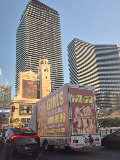The image depicts a street scene in Paradise, Nevada, USA, dominated by the Bellagio Hotel and Casino and a tall, modern building with a vertically striped facade. The camera is angled upwards. In the foreground, a white box truck with advertisements for a "GirlsDirectToYou.com" service is visible, featuring images of scantily clad women and a phone number. A black car with a California license plate reading "TOUSANT" is positioned behind the truck. Traffic lights and street signs are also visible.