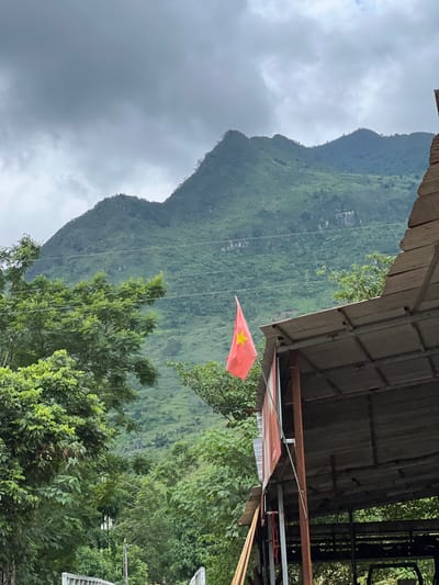 The image depicts a rural landscape in Lùng Tám Commune, Vietnam, characterized by lush green mountains under a cloudy sky. In the foreground, a rustic, open-air structure with a corrugated metal roof and weathered red siding is visible. A Vietnamese flag, red with a yellow star, flutters from a pole attached to the structure. Dense green foliage, including trees and shrubs, surrounds the building and extends up the slopes of the mountains. Power lines crisscross the scene, indicating some level of infrastructure. The overall atmosphere is one of natural beauty and simple living.