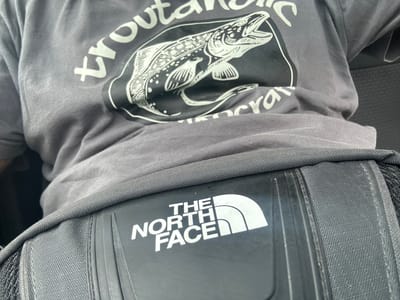 The image shows a close-up view of a person wearing a gray t-shirt with a graphic of a fish and text. Below the t-shirt, a black fanny pack or waist bag with "THE NORTH FACE" logo is visible. The fabric of the bag has a textured pattern. The background is out of focus, but appears to be an indoor setting with a dark textured surface. There are no visible indicators of the location in Starosel, Bulgaria.