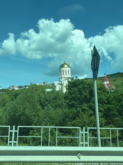 This image, taken in Varvarovka, Russia, depicts a landscape featuring religious architecture and natural elements under a bright blue sky with scattered white cumulus clouds.
In the foreground, a silver-colored metal road guardrail extends horizontally across the bottom, with a lighter gray metal fence featuring a grid-like design positioned above it. To the right, a gray pole for a road sign angles upwards and to the left, partially obscuring the scene beyond.
The midground is dominated by a dense expanse of lush green deciduous trees and bushes. Partially visible above these trees and to the left of the sign pole is a large, white or light-colored Orthodox church. This church features a prominent golden dome topped with a golden Orthodox cross, along with arched windows on its drum and walls. Further to the right and slightly in the background, partially hidden by the sign pole and trees, is a second building, also likely a church or chapel, characterized by a distinct red pyramidal roof and a smaller golden dome with a golden cross.
In the background, verdant green hills rise, with several modern-style houses or buildings with light facades and dark roofs visible on the left hill. No individuals are present in the scene. The overall impression is a sunny daytime view of a hilly, green environment.