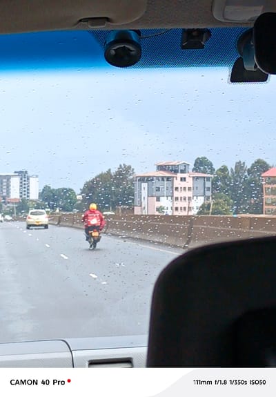 The image depicts a view from inside a vehicle looking through a rain-splattered windshield onto a multi-lane roadway in Gitaru ward, Kenya. A dark asphalt road with visible white lane markings appears wet, suggesting recent precipitation.
In the midground, a lone individual on a motorcycle, wearing a red jacket and a yellow helmet, is traveling away from the camera in the center lane. A red object, possibly a delivery bag, is secured to the rear of the motorcycle. Further ahead in the left lane, a light-colored sedan also moves away from the camera. A concrete road barrier runs parallel to the roadway, separating it from the background.
The background reveals an urban or suburban landscape with several multi-story buildings and interspersed green trees. A prominent light pink/beige building with grey sections and a multi-level roof structure is visible on the right, while a taller, grey building stands further to the left. The sky is overcast.
In the foreground, within the observing vehicle, a beige overhead console/visor area is visible. A black dashcam is mounted on the windshield, slightly left of center, with another smaller dark device to its right, and a partial rearview mirror on the far right. The top edge of a dark dashboard and the upper portion of a dark seat headrest are visible in the lower foreground.