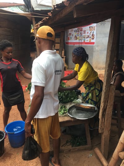 This vibrant image depicts a bustling outdoor market stall in Enyibichiri, Nigeria, under natural daylight. The setting is characterized by a rustic, open-air structure constructed from rough-hewn wooden beams and posts, with a dirt ground.
In the mid-ground, a woman wearing a bright yellow top, a traditional patterned wrapper, and a blue patterned headscarf is actively engaged in sorting or preparing large piles of fresh green leafy vegetables on a low counter or table. Beside the greens, a silver tray with a dark, rectangular object, possibly a weight, rests on the counter. Behind her, partially obscured by wooden beams, another person in a light-colored top is visible.
In the foreground, a man wearing a white t-shirt, a yellow baseball cap, and yellow shorts stands, holding a black plastic bag and a mobile phone. Near his feet, a blue plastic bucket, partially filled with water, sits on the ground. Another individual, wearing a black and red "Sports" branded top, is partially visible near the man, facing the bucket.
A white poster is affixed to a wall, showcasing a variety of food items, including meats, fish, and baked goods. Visible text on the poster reads "lotto: Customer Satisfaction is on priority," "SHOP B ENTRA," and "MARKET OPPOSITE FRUIT JUNCTIONISHIER." Other text fragments like "GROUP NIG" and "The Standard." are also discernable. The scene captures a moment of daily commerce and interaction within a local marketplace.