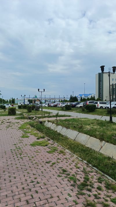 This media file depicts an outdoor scene in Barabinsk, Russia. The foreground features a brick pathway with overgrown weeds and a concrete drainage ditch. Beyond the path, a paved road leads to a parking lot filled with various cars. In the middle ground, there is a covered structure with a blue roof, possibly a bus stop or a small station, with a green-domed building visible behind it. Streetlights stand along the road. In the background, a modern multi-story building with a light green facade and white trim dominates the right side of the frame. Other buildings with blue roofs are also present. The sky is overcast with clouds, suggesting a cloudy or possibly rainy day. There are no visible people actively engaged in activities, though a few cars are parked, implying people have arrived or are about to depart. The overall impression is a somewhat unkempt but functional public space under a muted sky.