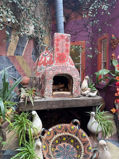 A whimsically decorated outdoor oven dominates the scene, adorned with red floral and swirling designs. It's mounted on a rustic wooden structure alongside ceramic dove figures and a large, patterned plate. Potted plants with long, green leaves add natural touches. The background features a purple building with orange trim and scattered decorative objects. The ground is covered in a tiled pattern of red, yellow, black, and grey. The entire scene has a quirky and artistic ambiance.
