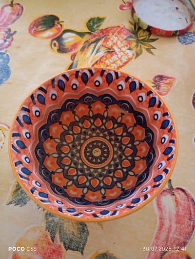 The image shows a close-up, overhead view of a decorative bowl with a mandala-like pattern in shades of orange, blue, and gray. The bowl is placed on a tablecloth with a vibrant, colorful print of fruits and vegetables, including what appear to be peppers, pineapples, and other produce. In the upper right corner, a partially visible metal lid with a similar fruit and vegetable design is present. The bottom left of the image shows the text "POCO C65", and the bottom right displays the date and time "30.07.2026 12:41". No people are visible in the image. The setting appears to be an indoor surface, likely a table, covered by the patterned tablecloth. There is no information in the image that suggests the location is in the city of Beruniy, Uzbekistan.