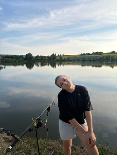 A young woman with blonde hair pulled back poses by a lake with fishing rods in the foreground. She is wearing a black t-shirt and light blue denim shorts. The lake reflects the sky and the distant treeline. The background features rolling hills with fields of sunflowers under a partly cloudy sky. The overall scene suggests a relaxed outdoor setting, likely a fishing spot in Tătărăști, Moldova.