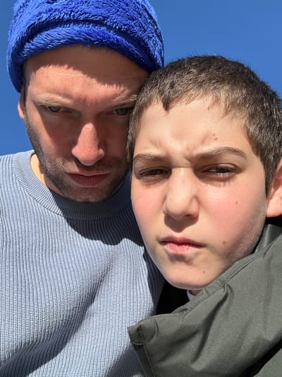An adult male and a younger individual are depicted in a close-up outdoor portrait, situated in Lipomo, Italy. The male, positioned on the left, wears a bright blue textured beanie and a light blue ribbed knit sweater. He has light stubble and a serious, direct gaze with furrowed brows and narrowed eyes. Strong, direct sunlight from the upper right illuminates his face, creating a pronounced shadow on his left side and torso. The younger individual, on the right and partially overlapping the male, has short brown hair and wears a dark olive green jacket over a visible white collar. Their expression mirrors the male's, featuring furrowed brows, narrowed eyes, and a slightly crinkled nose. Both subjects look directly into the camera. The background consists solely of a clear, bright blue sky, confirming an outdoor setting under direct sun. The close proximity of the individuals suggests an intimate composition.