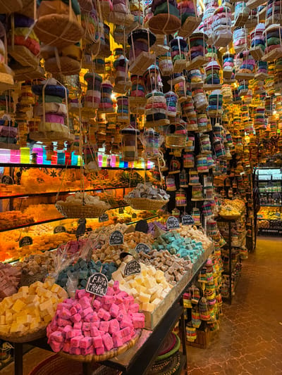 This image depicts the interior of a brightly lit market stall or shop, likely in Chefchaouen, Morocco, specializing in artisanal goods, specifically soaps. The scene is dominated by a dense display of colorful merchandise.
The ceiling is entirely covered with hundreds of individual soap products, each typically cylindrical or multi-layered, wrapped in diverse colored fabrics or papers, and suspended by twine in bundles of three to five items. Many bundles are also placed within small, shallow, light-colored baskets, which are then hung. The overall effect creates a canopy of vibrant, stacked forms extending throughout the visible space.
Below the suspended items, the shop features multi-tiered display tables and shelves. The foreground presents large, mounded piles of cubed or irregularly shaped soaps in various colors, including bright pink, light yellow, teal, light blue, brown, cream, and purple. These piles are arranged on metal trays and within woven baskets. Several white, handwritten labels indicate scents or types, such as "FRUTA TROPICAL" (Tropical Fruit), "MUSC BLANCO" (White Musk), "JASMIN CON LIMON" (Jasmine with Lemon), "HERB. LOUISA" (Louisa Herb), "MENTE ORIENTAL" (Oriental Mint), "MIEL CITRON" (Honey Lemon), and "PASSION FRUIT".
In the background to the left, further shelves are visible, densely packed with more uniformly shaped, yellowish-orange items, possibly additional soaps, which are brightly illuminated from behind, casting a warm glow. Above these shelves, horizontal panels of colored glass or plastic in blue, green, pink, and yellow are present, acting as decorative elements or light diffusers. To the right, a narrow aisle recedes into the distance, revealing more shelving units laden with products and a dark, vertical refrigeration or display unit at the far end.
The floor is a reddish-brown tile with a textured, possibly cracked or patterned, finish. No individuals are visible within the shop, and no direct actions or interactions are depicted. The lighting is predominantly artificial and warm, emphasizing the colors of the products.