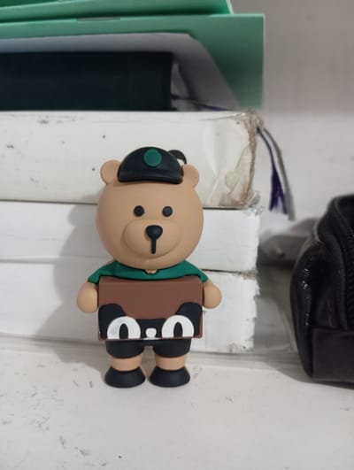 A close-up, eye-level shot shows a small, tan-colored teddy bear figurine with black eyes and a black nose, wearing a green collar and a brown shirt with white cartoon eyes. The figurine has a black cap with a green circle on top. It is positioned in the foreground against a background of stacked white books and a green folder. To the right of the figurine, a portion of a black zippered bag is visible. The scene appears to be indoors, on a shelf or table, with a white surface in the foreground. The overall lighting is soft, with shadows cast by the figurine and the objects behind it. The location is identified as Ак-Жар, Kyrgyzstan.