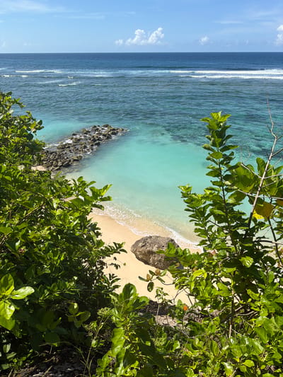 The image depicts a tropical beach in Benoa, Indonesia. The viewpoint is from an elevated position, looking down at the shoreline. Lush, green vegetation frames the right and lower portion of the image. The beach is a light sand color, with gentle waves lapping at the shore. The ocean transitions from a light turquoise near the beach to a deeper blue further out. A rocky outcrop extends into the water near the left side of the beach. The sky is visible on the left, with a few scattered clouds.