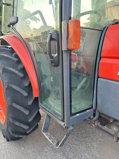 The image shows a close-up, eye-level, outdoor, medium shot of the side of a red and gray tractor. The tractor has large, black tires with deep treads. A cab door with a clear window is partially open, revealing a dark interior with some visible controls and possibly a seat. An orange rectangular light is mounted on a metal bracket to the left of the cab door. A metal step is attached below the cab door. The tractor is parked on a paved surface, likely a road or driveway, with some gravel visible. The background is blurred but suggests an outdoor environment with some greenery and structures, possibly residential buildings, indicating the tractor is located in Kableshkovo, Bulgaria. There are no people or animals present in the image. The lighting suggests it is daytime, possibly late afternoon, with sunlight reflecting off the tractor's surfaces and through the cab window.