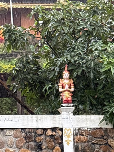 In the city of Ban Paem Bok, Thailand, an outdoor scene depicts a polychrome statue set against dense foliage and a building during rainfall. A low wall composed of rough, irregular dark brown and grey stones forms the base, topped by a weathered white concrete band. A white concrete pillar with a gold, stylized motif is visible on the right. Centrally, atop a white pedestal integrated into the wall, a light-skinned statue with a red and gold pointed headdress and ornate red and gold attire is depicted with hands pressed together in a gesture of reverence. Behind the wall and statue, dense green foliage, appearing wet, occupies the mid-to-background. In the upper background, a building with a dark roof featuring a prominent golden, serrated decorative trim is partially visible through the foliage. Rain is actively falling, evident from visible streaks in the air and wet surfaces.