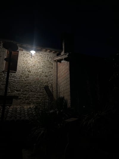The image depicts a nocturnal scene of an exterior stone wall with a lit lamp overhead. A dark tiled roof is visible in the foreground, and the background is predominantly black, suggesting darkness. There is a window on the stone wall, and part of a brick structure is visible to the right. Several plants are present in the lower right portion of the image, partially illuminated by the lamp. The scene is captured at night, possibly in a rural or older urban setting given the architectural style. The specific location in Cannara, Italy, cannot be definitively determined from the visual information provided.