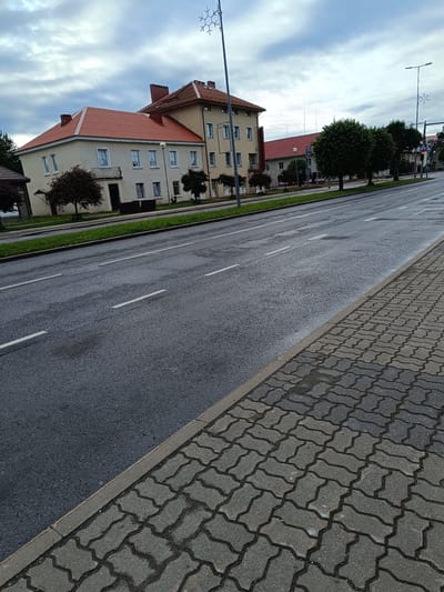 The image shows a street in Jõhvi, Estonia, on an overcast day. The street is paved with asphalt and marked with white lane lines. To the right of the street is a sidewalk paved with interlocking concrete pavers. The buildings along the street appear to be multi-story structures with red-tiled roofs. There are trees and streetlights visible, including one with a snowflake-shaped decoration. The overall scene suggests a typical urban environment in Jõhvi.
