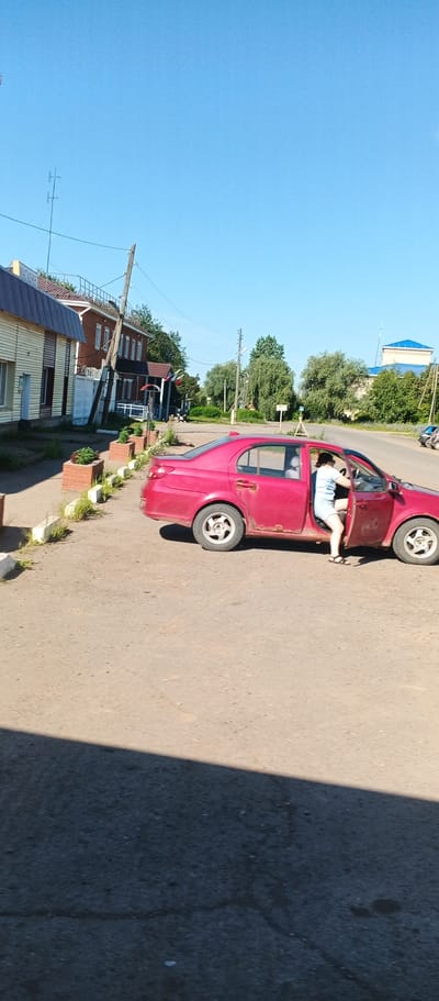 A full-shot vertical image depicts a red sedan with its driver's side door open. A person wearing a light-colored top and shorts is exiting the vehicle, stepping onto the asphalt road. The car is parked on the side of a rural road, with buildings lining the left side of the frame. These buildings appear to be commercial establishments, with one exhibiting a sign that reads "МАГАЗИН" (shop). The right side of the road is lined with trees. The sky is clear and blue, and the scene is bathed in sunlight, casting a shadow across the lower portion of the image. The location is identified as Kiyasovo, Russia.