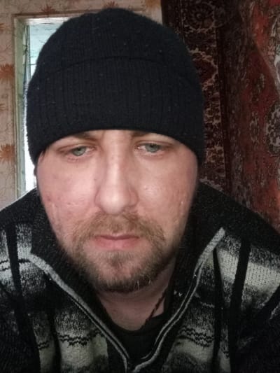 A close-up headshot of a middle-aged man with light blue eyes, a short beard, and a black beanie. He is wearing a black and white patterned sweater. The background consists of a doorway with a blurry green scene visible and a dark red patterned textile hanging. Based on the prompt, the location is Parabel, Russia.