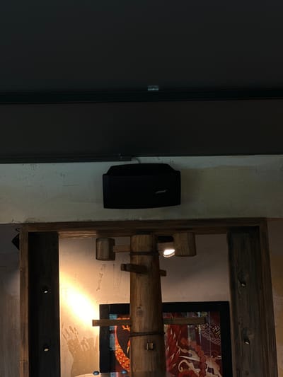 The image depicts an interior scene, possibly a room in Bonn, Germany, characterized by a rustic and slightly aged aesthetic. On the left, a large, dark grey door occupies a significant portion of the frame, its smooth surface contrasting with the textured wall to its right. Mounted on this wall is a black Bose speaker, its angular design hinting at its function.

To the right of the speaker, a wooden structure, seemingly a shelf or part of a decorative display, is prominently featured. This structure is constructed from rough-hewn, dark wooden beams, giving it a handcrafted and somewhat primitive appearance. Within this wooden framework, a bright light source illuminates the scene, casting a warm glow and creating highlights on the textured wall behind it.

A glimpse of artwork or a tapestry is visible within the wooden structure, displaying intricate patterns and vibrant colors, predominantly reds and oranges, with stylized figures that appear to be fantastical creatures or mythical beings. This artwork adds a touch of cultural or artistic interest to the otherwise minimalist and textured environment.

The lighting suggests that it is either evening or a dimly lit interior space. The overall mood is one of quiet ambiance, with a blend of modern technology (the speaker) and rustic charm. The rough textures of the wall and wood, combined with the warm light, create a cozy yet potentially artistic atmosphere. No people are visible in the image. The text "BOSE" is clearly visible on the speaker.