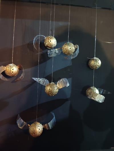 Seven golden-colored spherical objects are suspended against a dark, matte background in El Port De Sagunt, Spain. Each sphere, approximately 6 cm in diameter, features intricate geometric cut-out patterns. Transparent, irregularly shaped wing-like appendages, consistent with clear plastic film, are attached to the sides of each sphere. Each object is individually suspended by a thin, transparent filament extending vertically upwards. Illumination from the upper-left casts distinct shadows of the objects and their appendages onto the background. No individuals or active interactions are present in the scene.