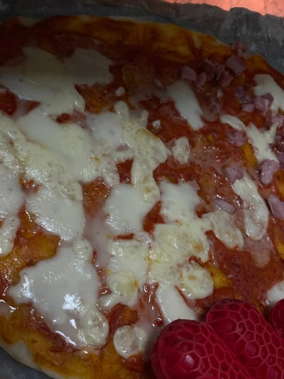 A close-up, overhead shot shows a pizza with melted cheese, tomato sauce, and pieces of ham. Two red, heart-shaped objects are visible in the lower right corner of the frame. The pizza appears to be on a dark surface, possibly a baking sheet or pan, with some of the edge visible. The lighting is dim, creating shadows on the pizza and obscuring some details. The location in Dumbrăvița, Romania is not discernible from the image.