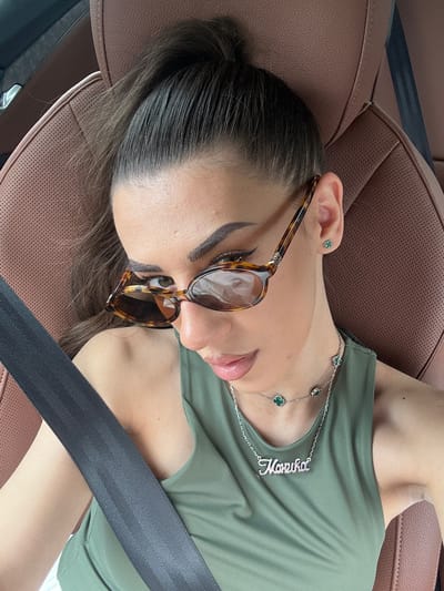 This media file features a close-up shot of a young woman reclining in a car seat. She is wearing a green t-shirt and a silver necklace with a nameplate that reads "Marika". She also has on tortoise-shell framed sunglasses and a small, green clover-shaped earring in her left ear. The car's interior is visible, with brown leather seats and a black seatbelt across her chest. The lighting suggests it is daytime. The woman's eyes are closed and her lips are slightly parted, giving the impression that she is resting or sleeping. The overall mood is relaxed and casual. The location is Studena, Bulgaria.