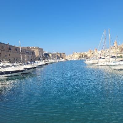 The image displays a marina scene located in Cospicua, Malta, within the Grand Harbour area, looking across a body of water towards the historic Three Cities. The foreground and middle ground feature a channel of blue-green water with subtle surface ripples. On the left, a substantial stone-built waterfront structure, appearing fortified and multi-storied with arched window apertures, runs along the shore. Moored parallel to this structure is a continuous line of approximately fifteen vessels, primarily white-hulled yachts and sailboats, with visible masts and rigging. On the right, further in the distance, another row of similar white-hulled boats is docked against a backdrop of a dense urban landscape. This cityscape is characterized by numerous light-colored historic buildings, prominent domes, and spires. The upper portion of the image consists of a clear, cloudless blue sky. No human figures or discernible activity are present in the scene.