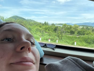 In the city of Van Hung, Vietnam, a close-up, low-angle selfie is taken from the perspective of someone lying down. The individual's face occupies the majority of the frame, with their forehead, eye, nose, and lips visible. Their left eye is open, showcasing blue-grey irises and long eyelashes. A light blue, textured object is positioned behind their head and to the left. To the right of the face, a pair of black sunglasses rests on a dark surface, possibly a window sill. In the background, a lush, green landscape unfolds with rolling hills and trees under a cloudy blue sky. Several red and white poles are visible along a track or road in the middle distance. The scene appears to be set within a vehicle, likely a train or bus, given the view through the window and the presence of what seems to be a seat or upholstery in the foreground.