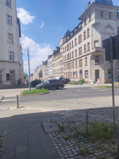 This is a street-level, outdoor, eye-level medium shot of a sunny day in Chemnitz, Germany. The scene depicts a quiet street lined with historic buildings on both sides, featuring an architectural style common in European cities. The buildings are multi-story, with many windows and some visible decorative elements.
On the left, a lighter-colored building with large windows dominates the foreground. Across the street, a row of taller, narrower buildings extends into the distance, with a more detailed building at the corner of the block in the mid-ground. The street is paved with cobblestones, and there's a crosswalk in the foreground. A dark-colored minivan is parked and driving on the street, along with other vehicles parked further down the road.
In the foreground, a person is walking across the street, and another person is walking on the sidewalk. The sky is bright blue with some fluffy white clouds, indicating fair weather. A street sign and a traffic light pole are visible. A washing machine sits outside one of the buildings in the mid-ground, a slightly unusual object in this context.
The time of day appears to be midday or early afternoon due to the bright sunlight and distinct shadows. The overall mood is calm and ordinary, capturing a slice of daily life in the city. There is no visible text that is legible.