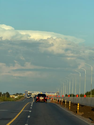 The image depicts a view from a vehicle traveling on a highway, likely leading into or out of Derzhavino, Russia, as suggested by the provided context. The road is paved with asphalt and marked with a solid yellow line on the left and dashed white lines on the right lane. Several cars are visible on the road, including a dark-colored sedan in the foreground, traveling away from the viewer. In the distance, a large structure, possibly a toll plaza or an overpass, is illuminated. The right side of the highway is bordered by a concrete barrier, with a row of streetlights and construction markers. The environment appears to be rural or suburban, with vegetation and some small buildings visible to the left of the road. The sky is partly cloudy, with dramatic formations of white and gray clouds set against a blue sky, suggesting either early morning or late afternoon.