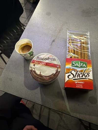 The image displays a table with a cup of espresso, a container of "Biscottata Mood" chocolate spread, and a package of "Salza Sticks" grissini. The objects are arranged on a textured surface, possibly a table in an outdoor seating area. The lighting suggests it is daytime, with shadows indicating sunlight. The background is blurred, showing what appears to be paving stones and dark, indistinct shapes. There are no people clearly visible in the frame, although a hand is partially visible in the lower right corner. The scene is forensic and matter-of-fact, documenting the presence and arrangement of these food items. The location of Zvornik, Bosnia and Herzegovina, is not discernible from the visual information provided in the image.