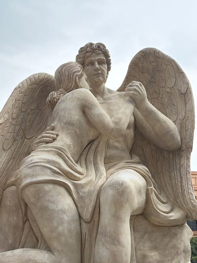 A low-angle, medium shot captures a large, light-colored stone sculpture of two winged figures embracing, set against an overcast sky. The figures, likely angels, are carved with detailed musculature and drapery. One figure, appearing more masculine, has outstretched wings and holds the hands of the other figure, who is positioned closer to the viewer, their back to the camera, and their head turned to face the first figure. The sculpture is located in Nghĩa Trụ Commune, Vietnam. The background shows a faint hint of a building with an orange facade to the right, suggesting an urban or park setting. The lighting is diffused, creating soft shadows and highlighting the texture of the stone.