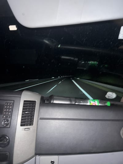 The image depicts the interior of a vehicle at night, looking out through the windshield at a highway. The dashboard is visible in the foreground, with control panels and an air vent on the left side. A bottle with a green cap and visible label is resting on the dashboard. The highway ahead is illuminated by streetlights and appears wet. There are no people visible in the image. The scene does not provide any information that could be used to determine its location in the city of Montreal, France.