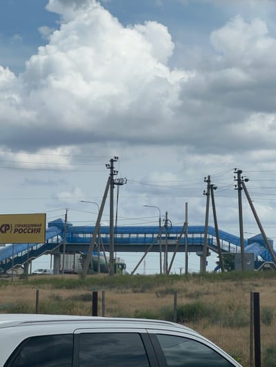The image shows a pedestrian overpass in the city of Prigorodny, Russia, under a cloudy sky. The overpass is blue and has ramps and stairs. A yellow sign with Russian text, including "СПРАВЕДЛИВАЯ РОССИЯ" (Fair Russia), is visible to the left of the overpass. Several utility poles with power lines are present, casting shadows on the scene. In the background, a white truck and a green truck are visible. The foreground shows the roof and window of a white vehicle, and a wire fence separates the road from a grassy area with dry vegetation. The overall scene appears to be a roadside view.