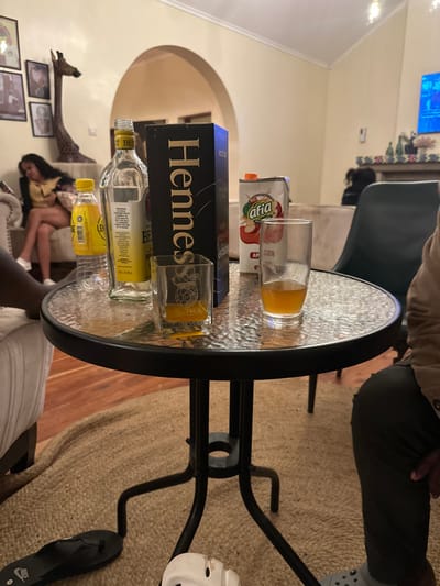 This image depicts an indoor residential scene, presumed to be within Olkaria ward, Kenya. The foreground is occupied by a round table with a black metal frame and a textured glass top. On the table, from left to right, are a clear plastic bottle of "BEST" branded lemonade, a largely empty clear glass bottle of "BEST DRY GIN 10 YEARS", a dark rectangular "Hennessy" box, a square glass containing a small amount of amber liquid and etched with "NOOR CUBA", a white "Afia" apple juice carton with a red cap, and a tall clear glass holding amber liquid, approximately one-third full.
Two individuals are partially visible in the immediate foreground, seated. On the left, a person's leg is visible, with a black Nike flip-flop resting on a round woven rug. On the right, a person's lower torso and leg are visible, wearing dark trousers and a grey Croc shoe. In the mid-ground left, a female individual with long dark hair, wearing a yellow top and light-colored shorts, is seated on a light-colored sofa, looking downwards with her legs crossed.
The room features light-colored walls, a wooden floor partially covered by a large circular woven rug, and light-colored sofas. A tall wooden giraffe statue stands against the back wall, which is adorned with several framed pictures. An arched doorway leads to an adjacent room. In the far background right, a wall-mounted television with a blue display is visible above a decorated mantelpiece. The scene is illuminated by dim, artificial indoor lighting, suggesting an evening or nighttime setting.