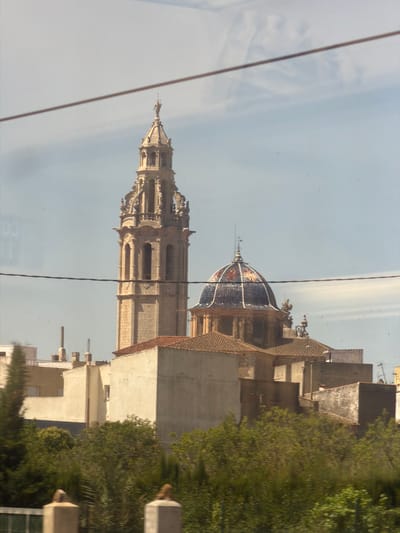 The image captures a view of the Church of Our Lady of the Assumption in Vinaròs, Spain. A prominent bell tower, characteristic of Gothic architecture, stands tall on the left side of the frame. It is intricately decorated with ornate carvings and features multiple arched windows. To the right of the tower, a colorful dome, adorned with blue and red tiles, crowns another section of the church. The buildings in the foreground are typical of the region, with terracotta-tiled roofs and light-colored walls. Lush green trees and foliage obscure the lower part of the scene, suggesting a sunny day with clear skies. The image appears to have been taken from a distance, possibly through a window, as evidenced by two horizontal lines that traverse the frame.