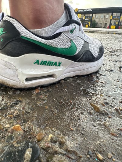 The image shows a close-up view of a person's foot wearing a Nike Air Max shoe, standing on a wet, gravelly surface, likely on a street or sidewalk. In the background, a gas station with the logo "КРАСНЕФТЬ" (Krasneft) is visible. The sky appears overcast. The overall scene suggests an outdoor urban environment.
