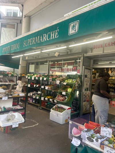This image depicts the entrance of a supermarket named "JIMMY ZHOU SUPERMARCHE" in Noisy-le-Grand, France. A prominent green awning stretches across the storefront, displaying the store's name and a logo. Another sign above the entrance reads "BOUN KHA," while a window banner advertises "PRODUITS EXOTIQUES."
Outside the store, a wide array of fresh fruits and vegetables is displayed on metal shelves and in green crates, resembling a street market stall. Visible produce includes watermelons, guavas, mangoes, papayas, green leafy vegetables, and yams. Several price labels are attached to the produce, showing items like "IGNAME 6 99" (Yam, 6.99), "FRUTA PAN 6 99" (Breadfruit, 6.99), "GOYAVE ROSE 9 99" (Pink Guava, 9.99), and "Proteque 3 80" (Watermelon, 3.80). Some labels also indicate origins, such as "ORIGINE REP DOMINOX" (Origin Dominican Republic) and "ORIGINE ALLEMAGNE" (Origin Germany), with a reference to "RUNGIS" (a major wholesale market).
Two individuals are present: one person wearing a dark top and light pants stands inside the store, appearing to work or attend to goods, and another person in a light hoodie is browsing the outdoor produce section. The scene takes place during the daytime, under bright, likely overcast, conditions. The image captures the lively, functional atmosphere of a neighborhood grocery store specializing in diverse and exotic produce.