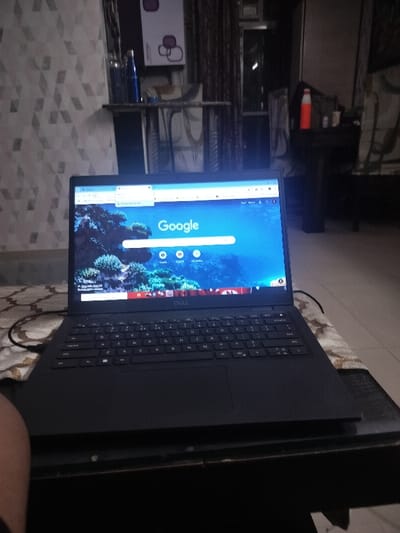 A black Dell laptop with a Google search page open on the screen is sitting on a brown table. The table is partially obscured by a person’s leg in the foreground. The laptop is connected to a black cord running off-screen. Behind the table, there are various out-of-focus objects, including furniture, a door, and a window. The scene appears to be set in a dimly lit room, possibly an office or home. The coordinates provided (28.577, 77.392) place the image in Noida, India.