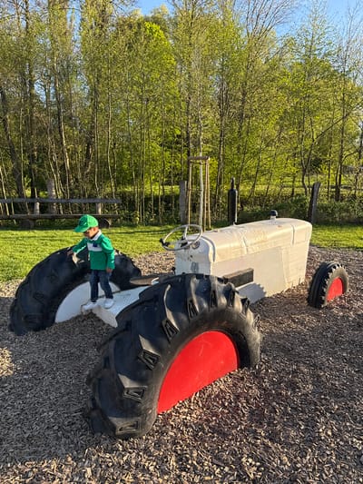 The image, taken in Olingen, Luxembourg, depicts a sunny outdoor playground scene. A young child, wearing a green long-sleeved shirt, blue jeans, white sneakers, and a green baseball cap, is standing on a large, white playground tractor. The child is positioned on the side of the tractor, between its body and one of its oversized black tires.
The tractor itself is a prominent feature, primarily white with large black tires that have red inner wheel detailing. A steering wheel is visible at the front of the tractor. Another loose tire, similar in appearance with black rubber and a red accent, lies on the wood chip ground near the tractor's rear. The ground throughout the visible area is covered in light-colored wood chips, commonly used for playground surfacing.
In the background, beyond a grassy area, a line of trees with vibrant green leaves suggests a spring or early summer setting. A wooden fence is also visible among the trees. The sky is clear and blue, indicating bright, sunny weather. The shadows cast by the tractor and the child point to either morning or afternoon sun. The brand name "Continental" is faintly visible on the side of one of the large black tires. The scene portrays a child engaged in play on the tractor in an inviting outdoor environment.