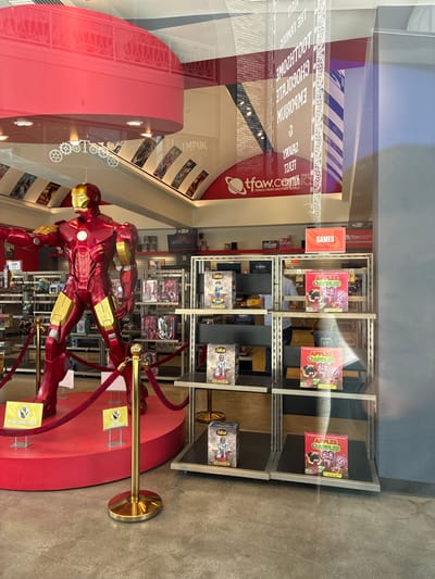 The image shows the interior of a comic book and collectibles store. A large, detailed replica of Iron Man stands prominently in the foreground, positioned on a red circular platform. The replica is made of shiny red and gold material, and is in a dynamic pose. Red velvet ropes and stanchions, with gold bases, surround the platform. Two yellow signs with black hand icons and the text "PLEASE DO NOT TOUCH" are visible on the platform.
Behind the Iron Man replica, shelves are filled with various merchandise. Some shelves display boxes of "Fallout" figures, and others hold boxes of "Apples to Apples" card games. The store appears well-lit, with overhead lighting visible. Reflections in the store window show some of the store's signage, including a red logo with "tfaw.com" and the tagline "THINGS FROM ANOTHER WORLD". There are also reflections of vertical stripes in blue and white on a pillar, and text that appears to be reversed.
In the background, more shelves of merchandise are visible, along with posters or displays on the walls. A glimpse of a person in a grey shirt is visible in the background between shelves, walking away from the camera. The floor is made of concrete. The overall impression is of a retail environment dedicated to pop culture and collectibles.
