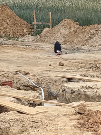 The media file displays a construction site in Opatów, Poland. The scene is dominated by rough, sandy terrain with some areas of green vegetation visible on the left. Several wooden planks are positioned vertically and diagonally, suggesting ongoing construction work.
In the central area of the image, a black rectangular object with some red accents is visible. This object appears to be a piece of equipment or machinery, possibly a generator or a portable power source, with a cable extending from it. A white cable or hose is also seen snaking across the sandy ground in the middle right of the frame.
The lighting suggests it is daytime, though the specific time is difficult to ascertain. The overall impression is one of an active construction project in a somewhat arid or disturbed landscape. There is no visible text in the image.